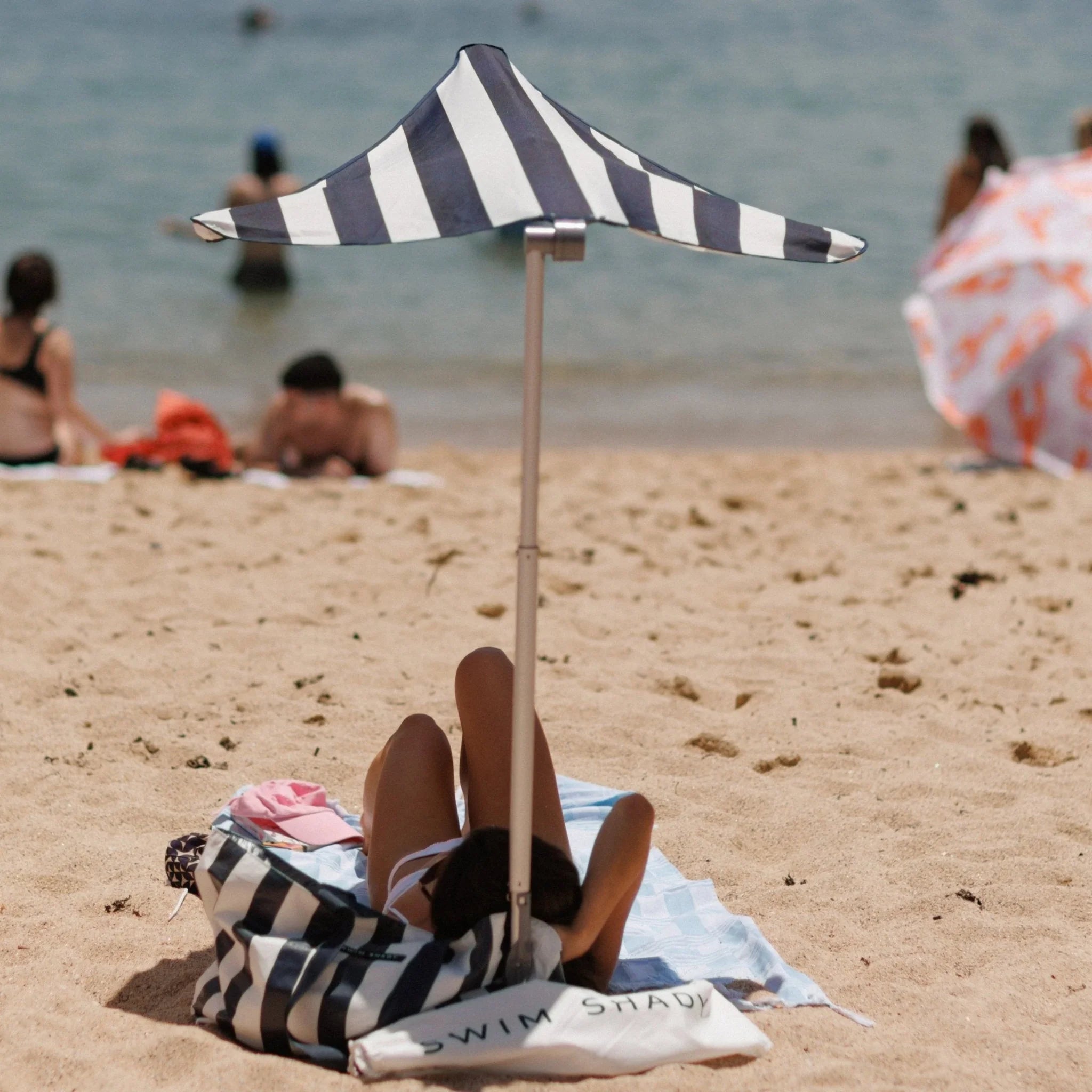 Person lying on a towel under a striped beach umbrella with people and ocean in the background