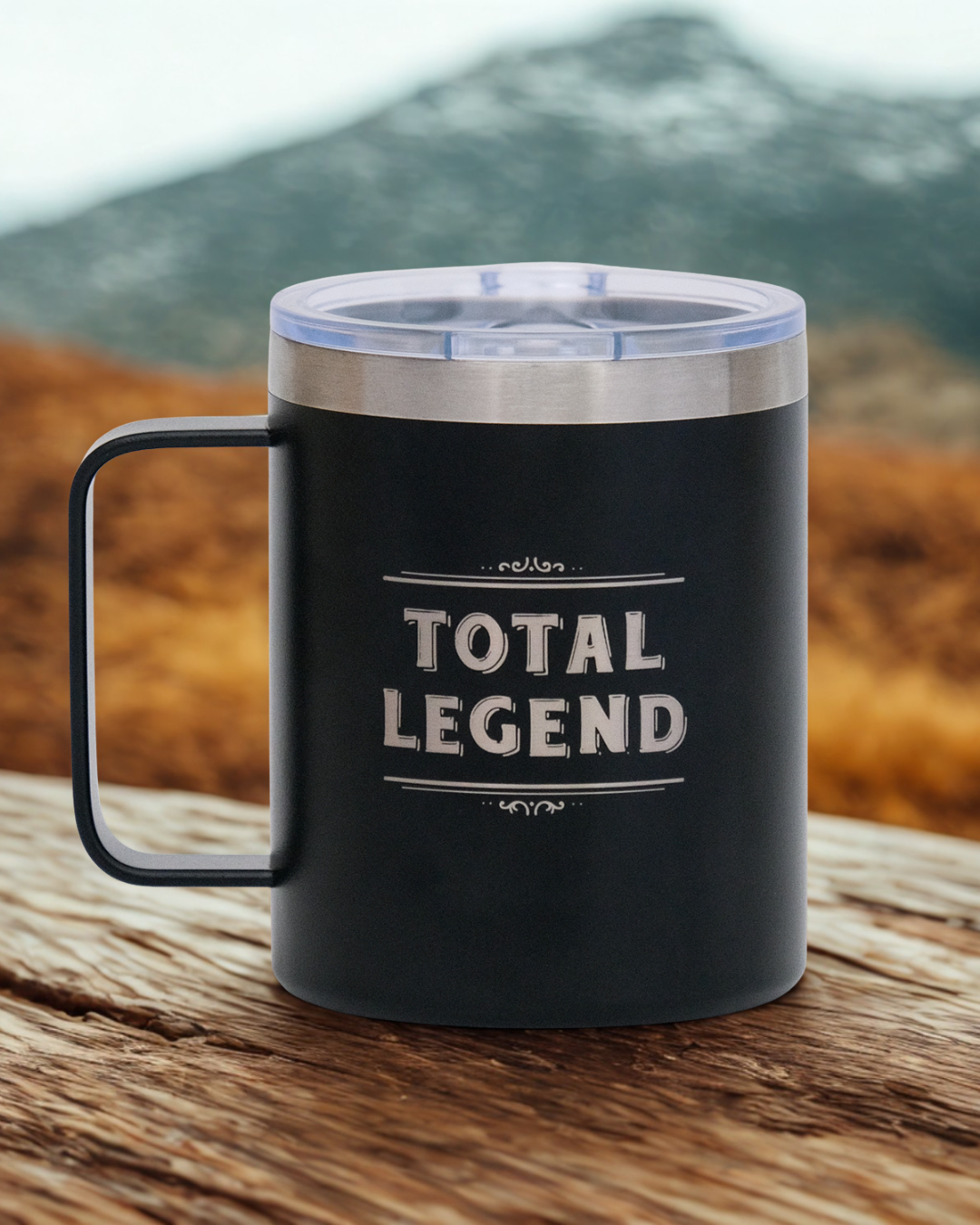 Black mug with 'TOTAL LEGEND' text on a wooden surface with a mountainous background