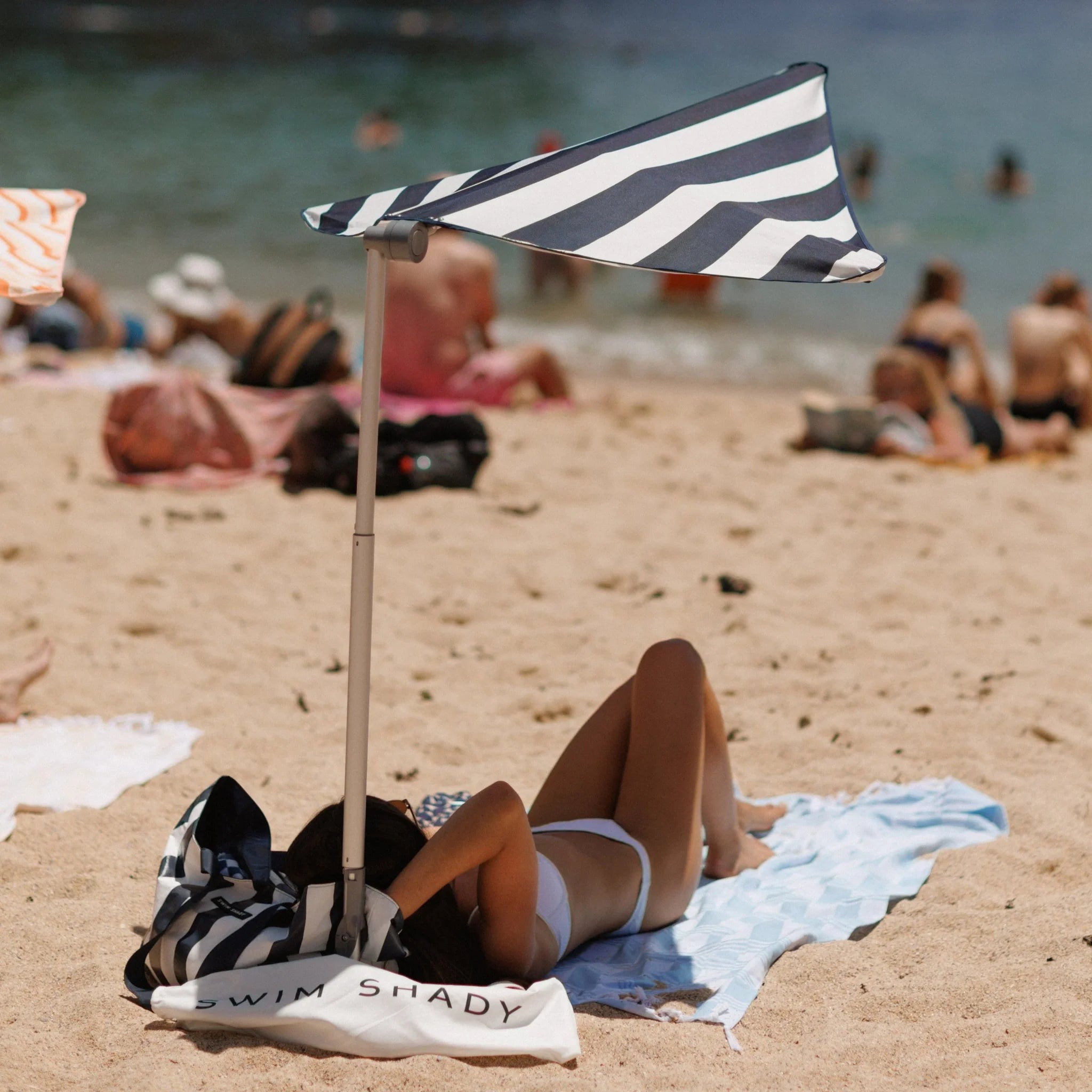 Person lying on a towel under a striped beach umbrella with 'Swim Shady' branding on a sandy beach.